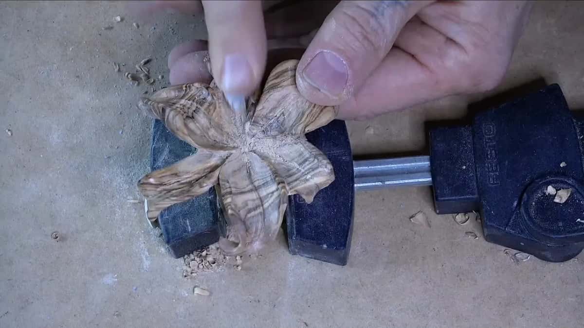 A small carved flower in heavily figured wood held between two black clamp jaws, fine dust around it.