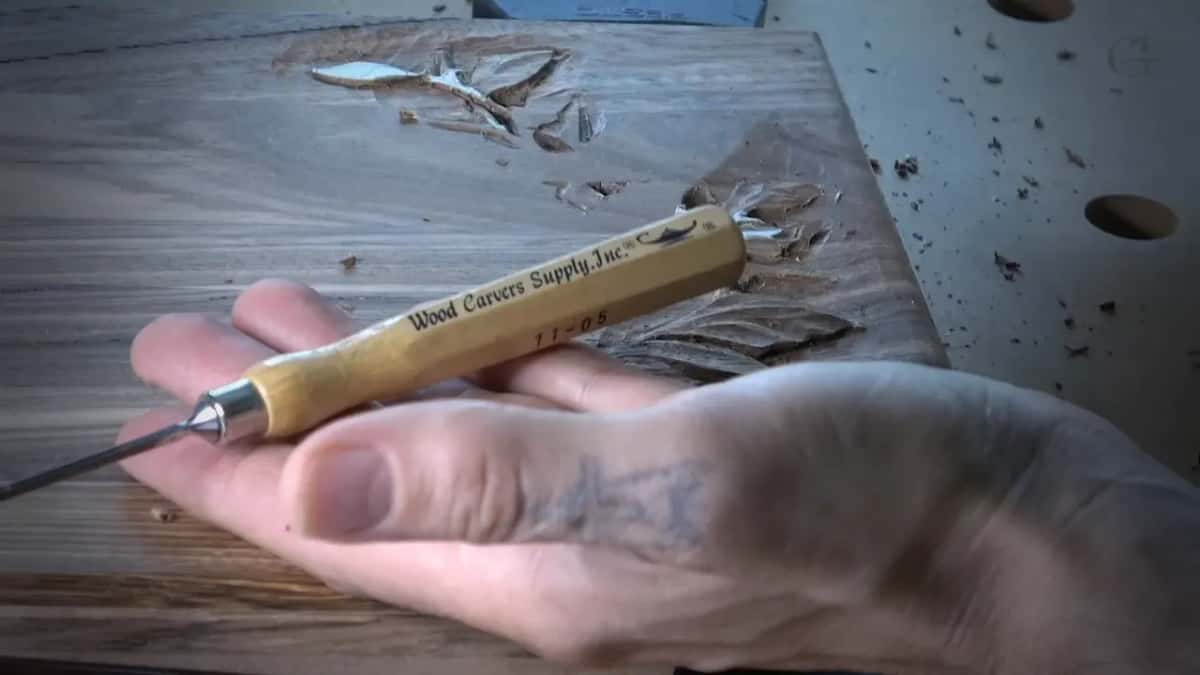 A hand holds up a gouge with its maker's name burned into the handle, above the dark roughed board.