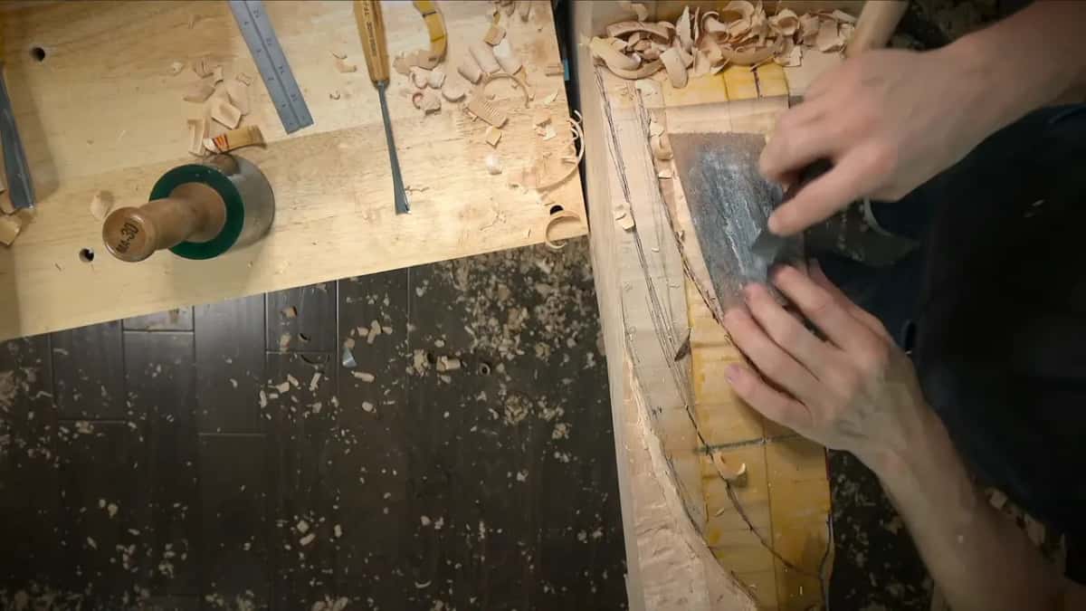 Two hands press a wide steel blade against a curved blank while gouges, a ruler and a mallet head wait on the bench.