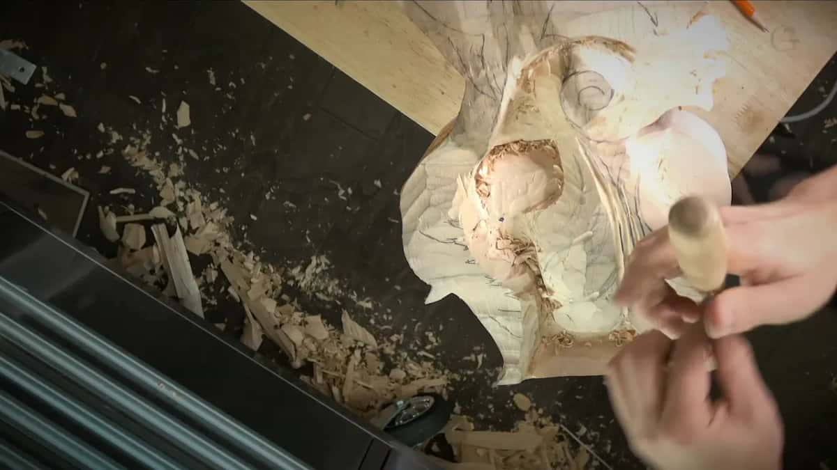 Overhead view of both hands on a gouge handle above a pale carved leaf panel, chips spread over the dark bench.