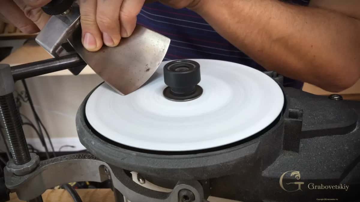 A wide fan-shaped blade held in a jig meets the spinning white honing wheel of a sharpening machine.