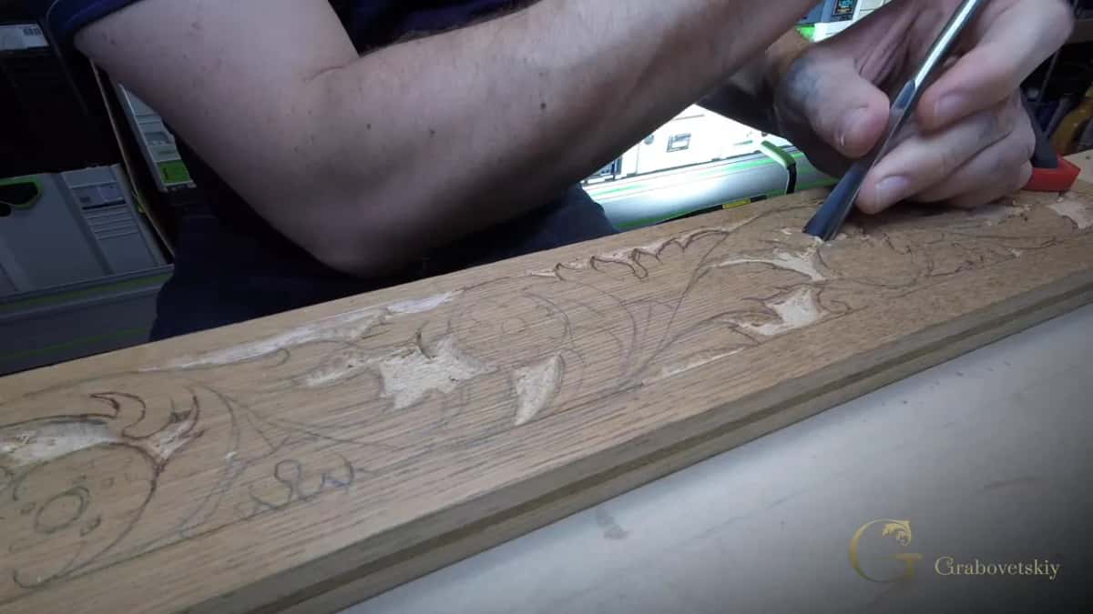 A long tan board with coarse open grain, half its leaf scroll carved and the rest still in pencil, chisel in hand.