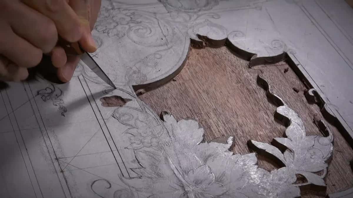 A knife lifts waste from a chalked design, opening a window of dark brown wood with straight, quiet grain.