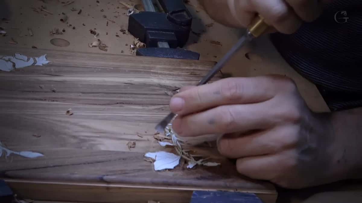 A small gouge carves a leaf into a brown board full of dark streaks, dry chips scattered over the surface.